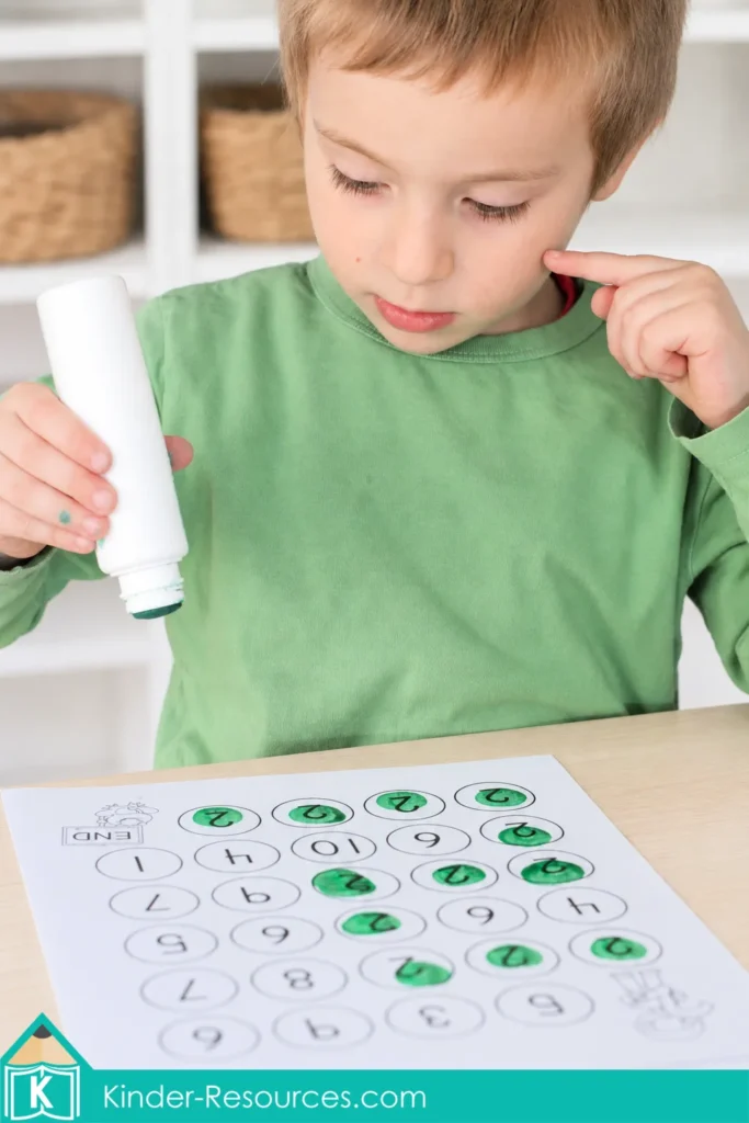 Preschool letter J math activity with a child using a green dot marker to find numeral 2 on a maze number recognition worksheet.