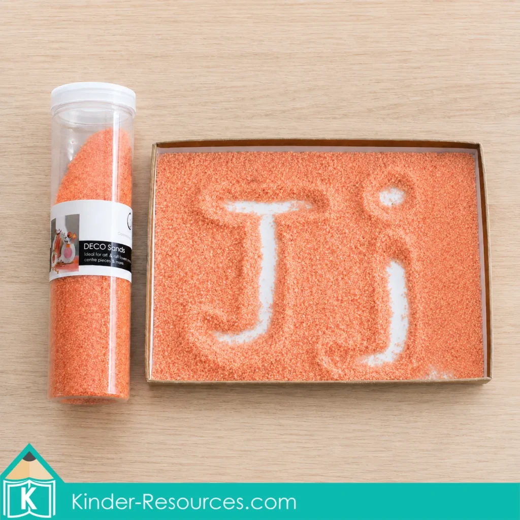 Preschool Letter J sand tray activity with uppercase and lowercase J traced in peachy orange sand beside a container of matching colored sand.