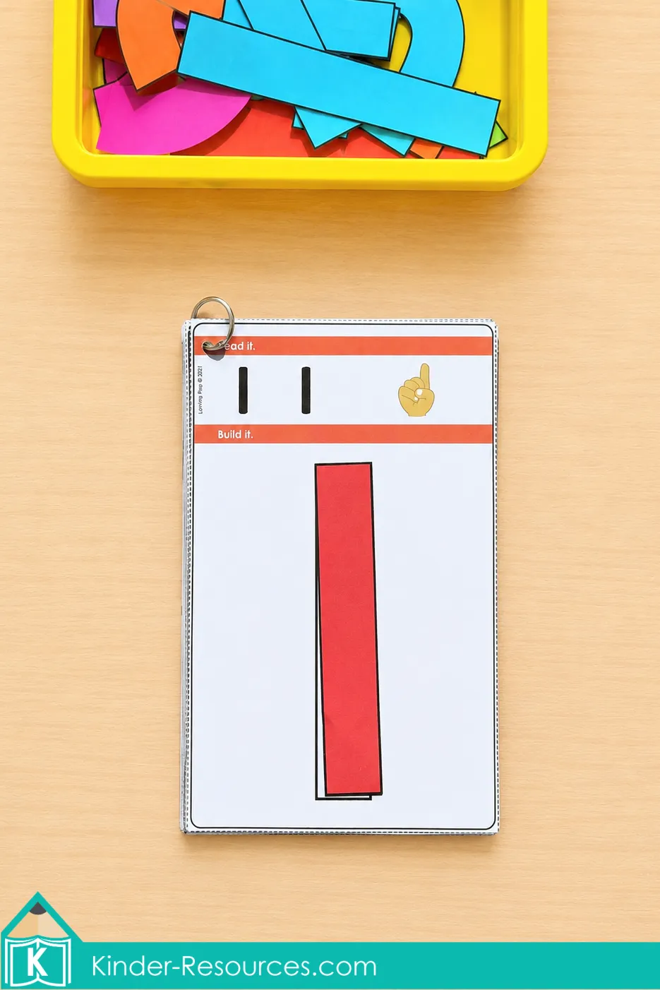 Using a red rectangular piece to build number 1 on a ring-bound activity card below a yellow tub.