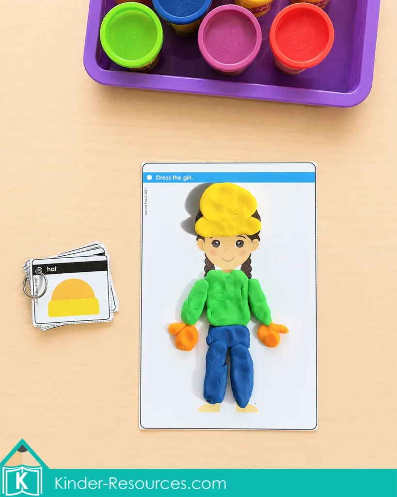 Using playdough to dress a printable girl in a hat, shirt, pants, and mittens beside clothing vocabulary cards.