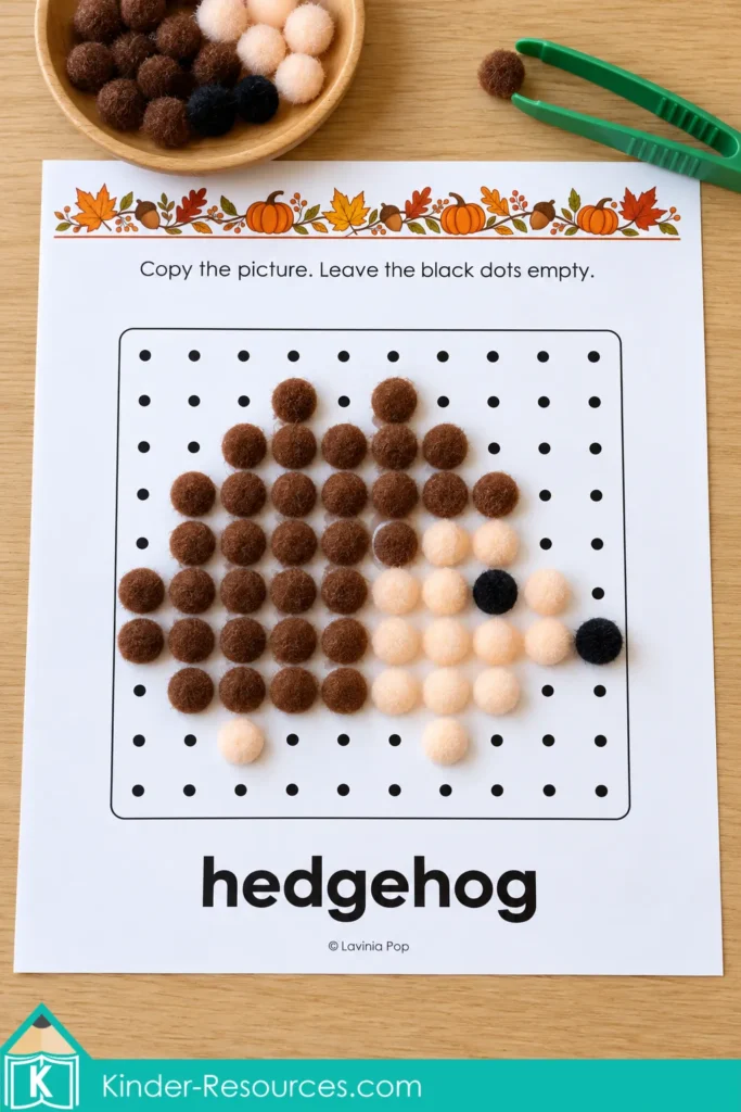 Brown, cream, and black pom-poms form a hedgehog on a printable dot-grid mat, with green tweezers and a bowl of pom-poms nearby.